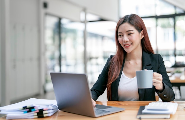 Joven empresaria sonriente en traje sosteniendo una taza y leyendo documentos en una computadora portátil en una oficina de espacio abierto