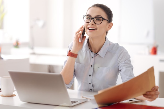 Joven empresaria sosteniendo el teléfono cerca de la oreja con gafas mientras está en el lugar de trabajo