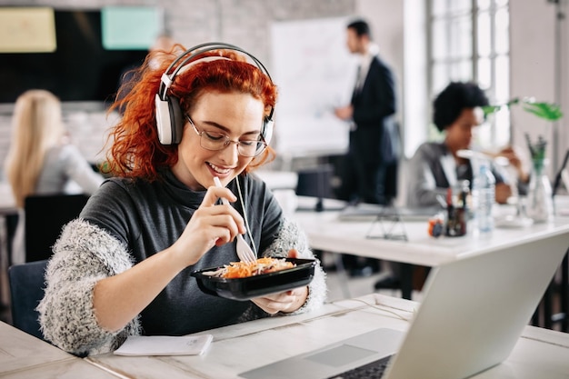 Joven empresaria tomando un descanso y comiendo ensalada mientras escucha música con auriculares en la oficina Hay gente en el fondo