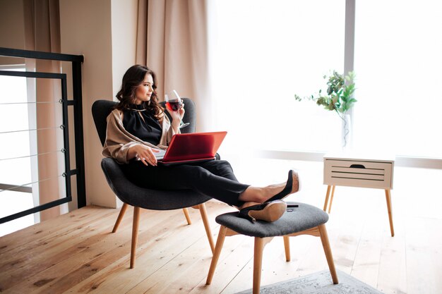 Joven empresaria trabajar en casa. Sentado en la silla y beber vino tinto de vidrio. Trabajo remoto. Portátil rojo en las rodillas. Solo en la sala. Luz.