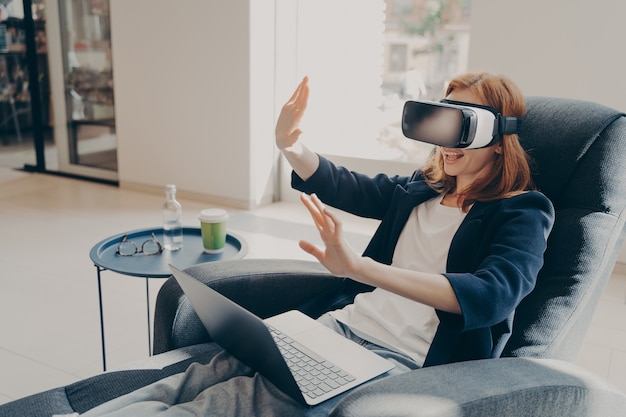 Joven empresaria vistiendo vr auriculares gafas y portátil sentado en un acogedor sillón en el vestíbulo de la oficina