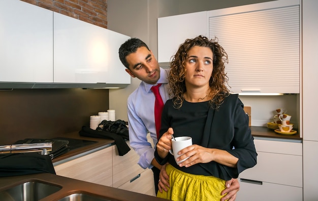 Joven empresario abrazando a la mujer rizada ofendida mientras sostiene una taza de café en la cocina