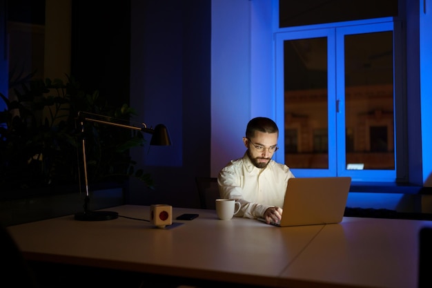 Joven empresario adicto al trabajo quedarse hasta tarde en el trabajo de oficina oscuro en la idea o estrategia de inicio en la computadora portátil