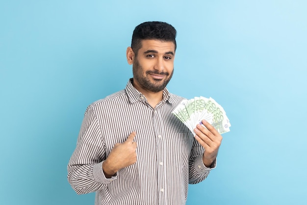 Joven empresario adulto con barba sosteniendo billetes de euro y apuntándose a sí mismo con orgullo