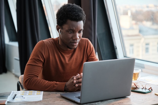 Joven empresario africano en auriculares sentado en la mesa y mirando el monitor de la computadora mientras está sentado en la oficina