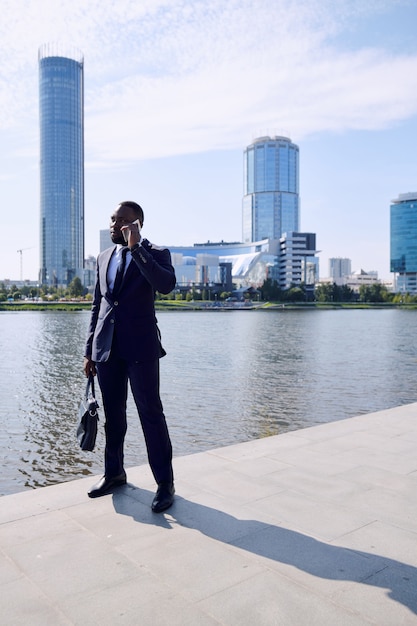 Joven empresario africano elegante o cliente consultor de agente en teléfono móvil mientras está de pie contra la orilla del río y el paisaje urbano con rascacielos