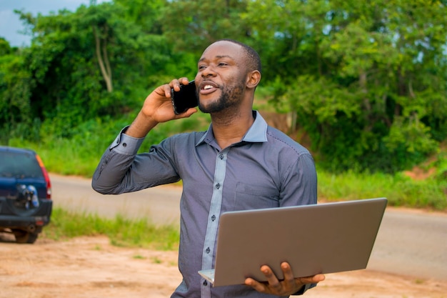 Joven empresario africano guapo de pie en una granja, sintiéndose sobreexcitado mientras sostiene su computadora portátil y hace llamadas con su teléfono celular.