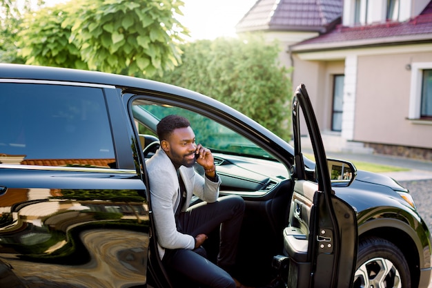 Joven empresario africano guapo sonriente sentado en el coche en el asiento del pasajero y hablando por teléfono. Construcción, casa y árboles verdes