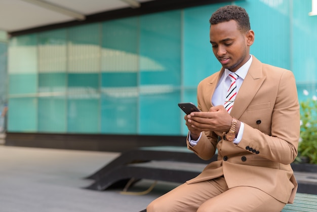 Joven empresario africano guapo con teléfono sentado fuera del edificio