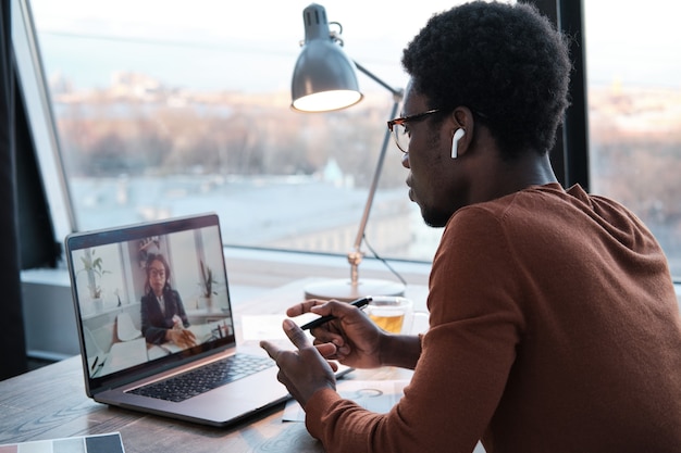 Joven empresario africano hablando con su colega en línea usando la computadora portátil mientras trabaja en la oficina