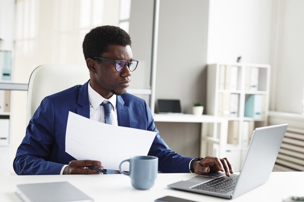 Joven empresario africano sentado en la mesa frente a la computadora portátil escribiendo documentos y haciendo trámites en la oficina