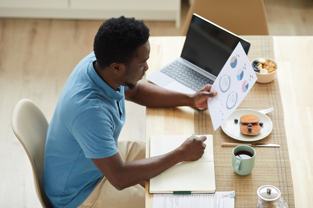 Joven empresario africano sentado en la mesa frente a la computadora portátil y examinar documentos durante su desayuno en casa