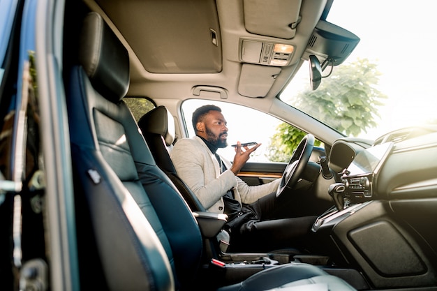 Joven empresario afroamericano exitoso hablando por altavoz a través del micrófono con el cliente, sentado en el coche caro. Negociaciones y reuniones de negocios.