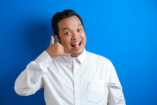 Joven empresario asiático en camisa blanca formal sonriendo a la cámara y muestra el gesto de signo de mano de teléfono llámame, contra el fondo azul.