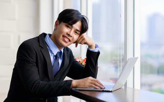 Joven empresario asiático es elegante y guapo con traje negro sentado mientras escribe en el teclado en la computadora portátil junto a la ventana en la oficina por la mañana.