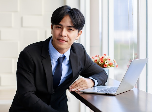 Joven empresario asiático es elegante y guapo con traje negro sentado mientras escribe en el teclado en la computadora portátil junto a la ventana en la oficina por la mañana.
