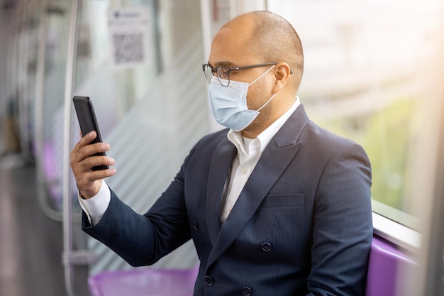 Joven empresario asiático con máscara de protección usando un teléfono inteligente sentado en skytrain. Empresario en viajes urbanos por la ciudad en tren aéreo.