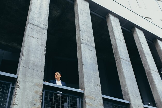 Joven empresario asiático de pie cerca del moderno edificio de oficinas negro de hormigón, en la terraza del balcón con columnas