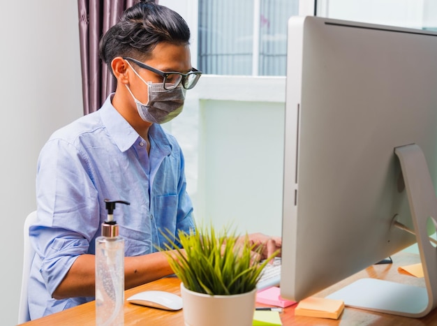 Un joven empresario asiático que usa una máscara protectora con gel desinfectante al lado de su trabajo desde la oficina en casa de forma remota en línea con la computadora PC pone en cuarentena la enfermedad coronavirus o COVID-19