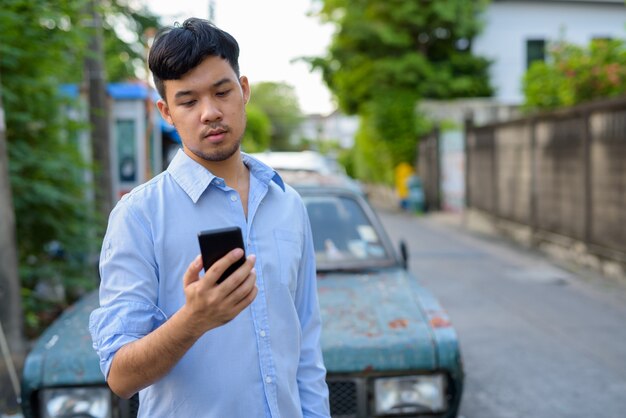 Joven empresario asiático mediante teléfono móvil contra coche viejo oxidado