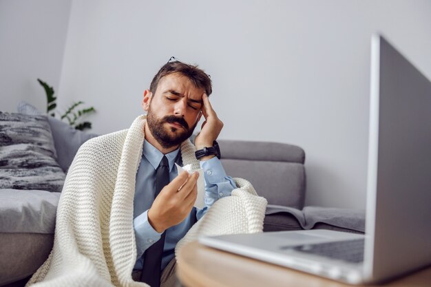 Joven empresario barbudo atractivo sentado en el suelo en casa y sosteniendo la cabeza porque tiene dolor de cabeza