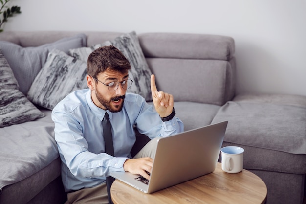 Joven empresario barbudo atractivo sentado en el suelo en casa, usando la computadora portátil y teniendo una idea de cómo resolver un problema.