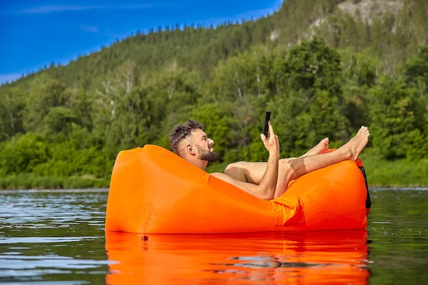 Joven empresario barbudo caucásico con teléfono móvil en la mano está acostado en una tumbona inflable naranja, que está nadando en el río cerca del bosque, ecoturismo.