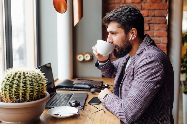 Joven empresario barbudo concentrado en auriculares usando laptop y bebiendo café con leche