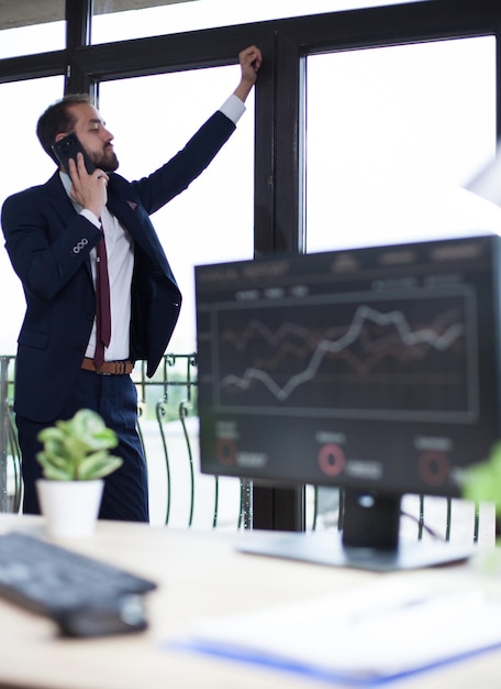 Joven empresario barbudo confiado mirando por la ventana y teniendo una conversación de negocios por teléfono. Emprendedor serio.