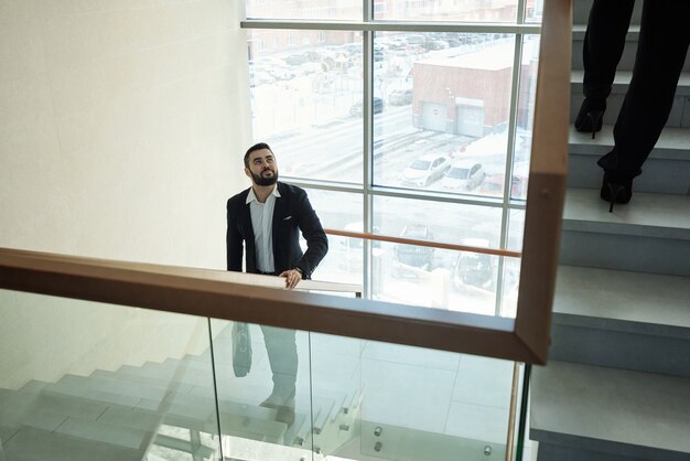 Joven empresario barbudo elegante con bolso de mano subiendo a su oficina dentro del centro de negocios contemporáneo