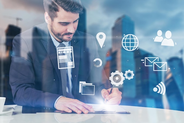 Joven empresario barbudo escribiendo en el portapapeles en la mesa de la oficina sentado sobre el fondo del paisaje urbano con doble exposición de la interfaz de Internet. Concepto de alta tecnología en los negocios. Desenfoque de imagen tonificada