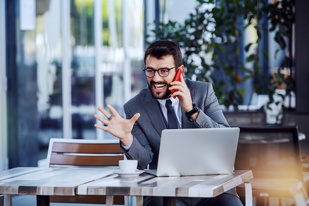 Joven empresario barbudo feliz caucásico en traje y con anteojos habiendo llamado por teléfono inteligente mientras está sentado en la cafetería. En la mesa hay café y computadora portátil.