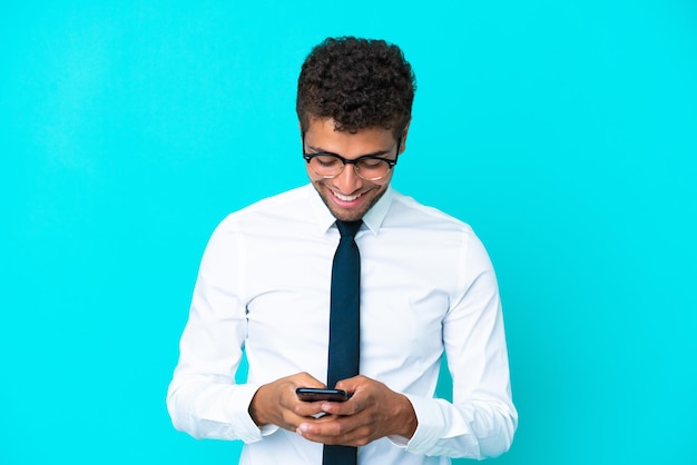 Joven empresario brasileño aislado de fondo azul enviando un mensaje con el móvil