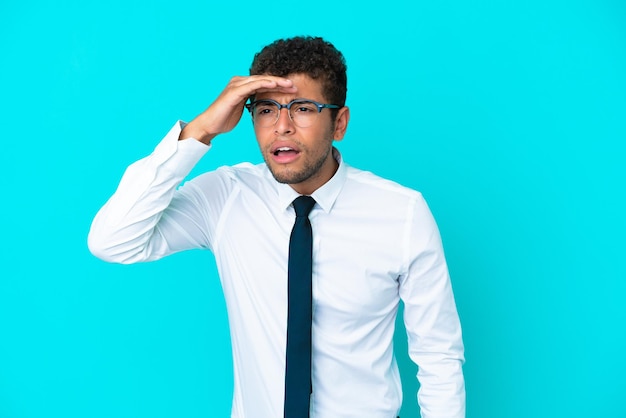 Joven empresario brasileño aislado de fondo azul mirando lejos con la mano para mirar algo