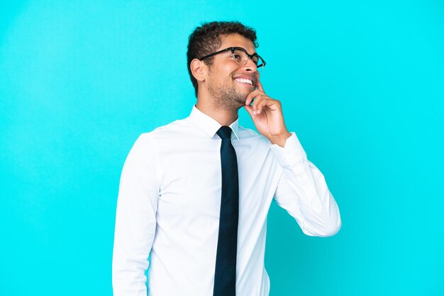 Joven empresario brasileño aislado de fondo azul pensando en una idea mientras mira hacia arriba