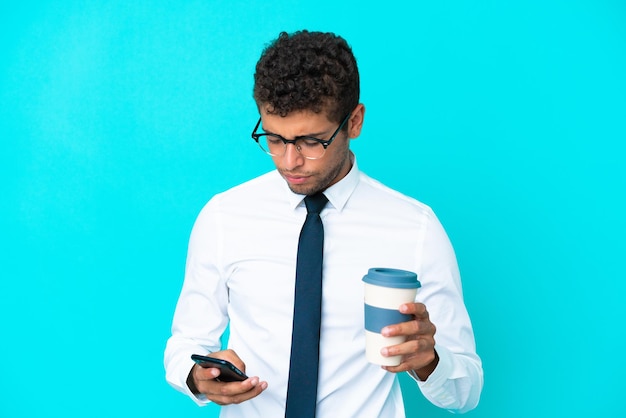 Joven empresario brasileño aislado sobre fondo azul con café para llevar y un móvil