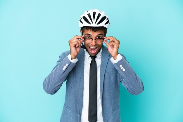 Joven empresario brasileño con casco de bicicleta aislado de fondo azul con gafas y sorprendido