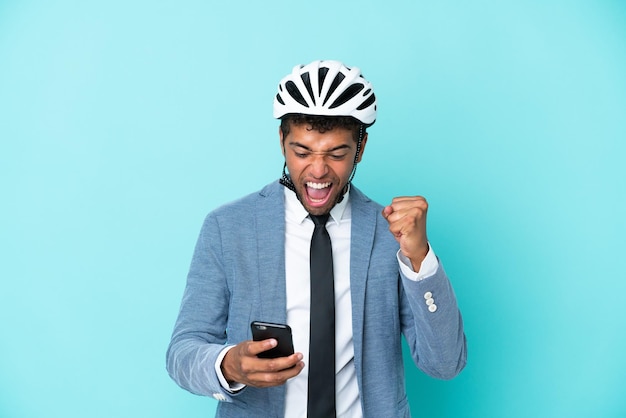 Joven empresario brasileño con casco de bicicleta aislado de fondo azul con teléfono en posición de victoria