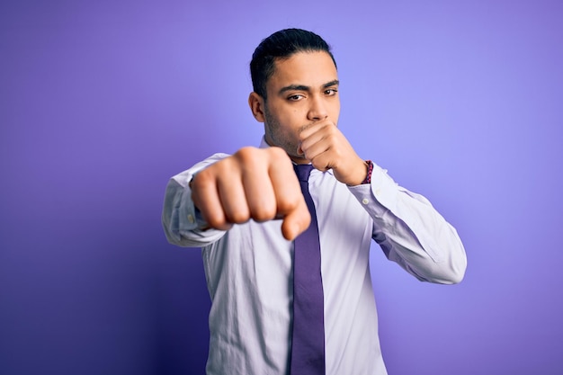 Joven empresario brasileño con elegante corbata de pie sobre un aislado fondo morado Golpeando el puño para luchar contra la amenaza y la violencia agresivas y enojadas