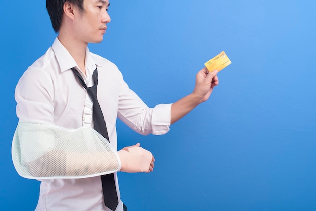 Un joven empresario con un brazo lesionado en un cabestrillo sosteniendo una tarjeta de crédito o tarjeta de seguro médico sobre fondo azul en el estudio, el seguro y el concepto de salud