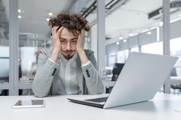 Un joven empresario con el cabello rizado parece estresado y cansado mientras trabaja en una computadora portátil en un moderno