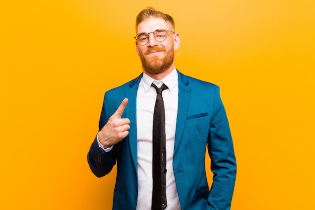 Joven empresario de cabeza roja mirando orgulloso, confiado y feliz, sonriendo y apuntando a sí mismo o haciendo el signo número uno