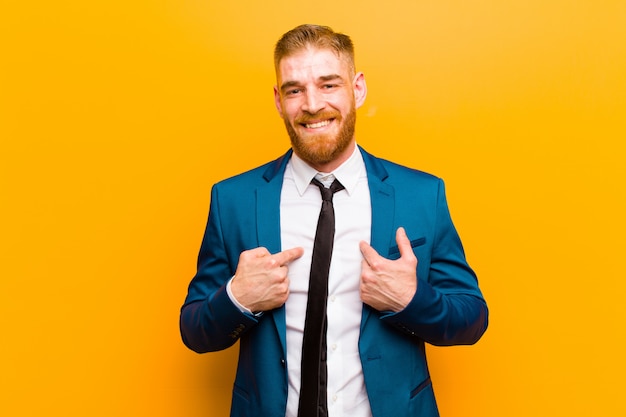 Joven empresario de cabeza roja se siente feliz sorprendido y orgulloso apuntando a sí mismo con una mirada emocionada asombrada sobre fondo naranja