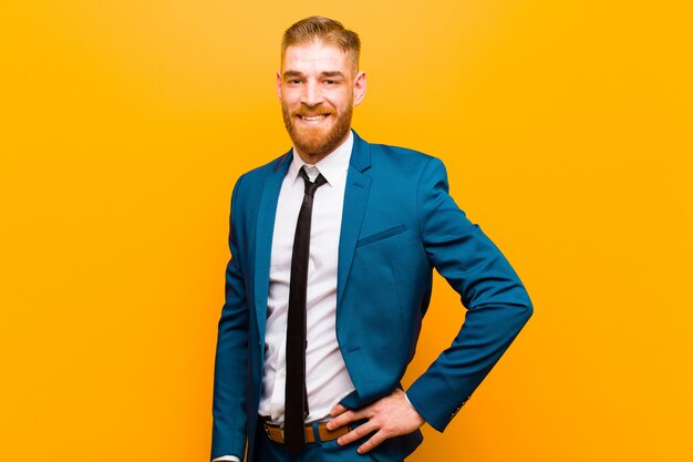 Joven empresario de cabeza roja sonriendo felizmente con una mano en la cadera y actitud segura, positiva, orgullosa y amigable