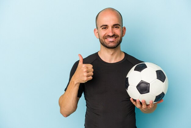 Joven empresario calvo jugando al fútbol aislado sobre fondo azul sonriendo y levantando el pulgar hacia arriba