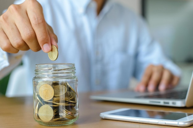 Joven empresario con camisa azul sosteniendo monedas poniendo en vidrio en el escritorio con espacio de copia Concepto de ahorro de dinero para la educación y la jubilación