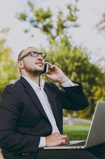 Joven empresario de camisa blanca, traje clásico, gafas. El hombre se sienta en un puf suave, trabaja en una computadora portátil, habla por teléfono móvil en el parque de la ciudad en el césped verde al aire libre. Oficina móvil, concepto de negocio.