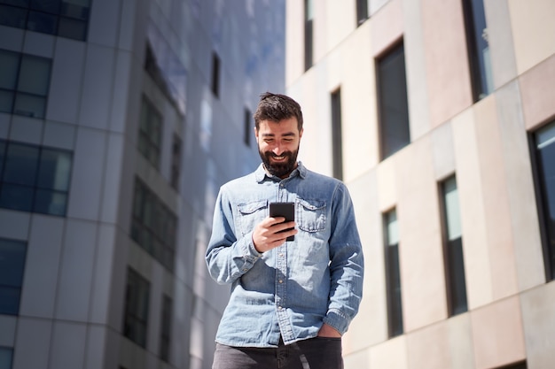 Joven empresario en camisa de mezclilla con teléfono inteligente cerca de edificios de oficinas en el centro financiero de la ciudad