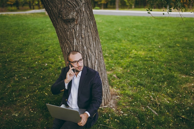 Joven empresario en camisa, traje, gafas. El hombre se sienta en el suelo de la hierba, habla el teléfono móvil om, trabaja en la computadora portátil de la PC en el parque de la ciudad en césped verde al aire libre en la naturaleza. Oficina móvil, concepto de negocio.