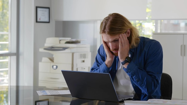 Joven empresario cansado que se siente estresado y abrumado sentado en una oficina moderna con una laptop